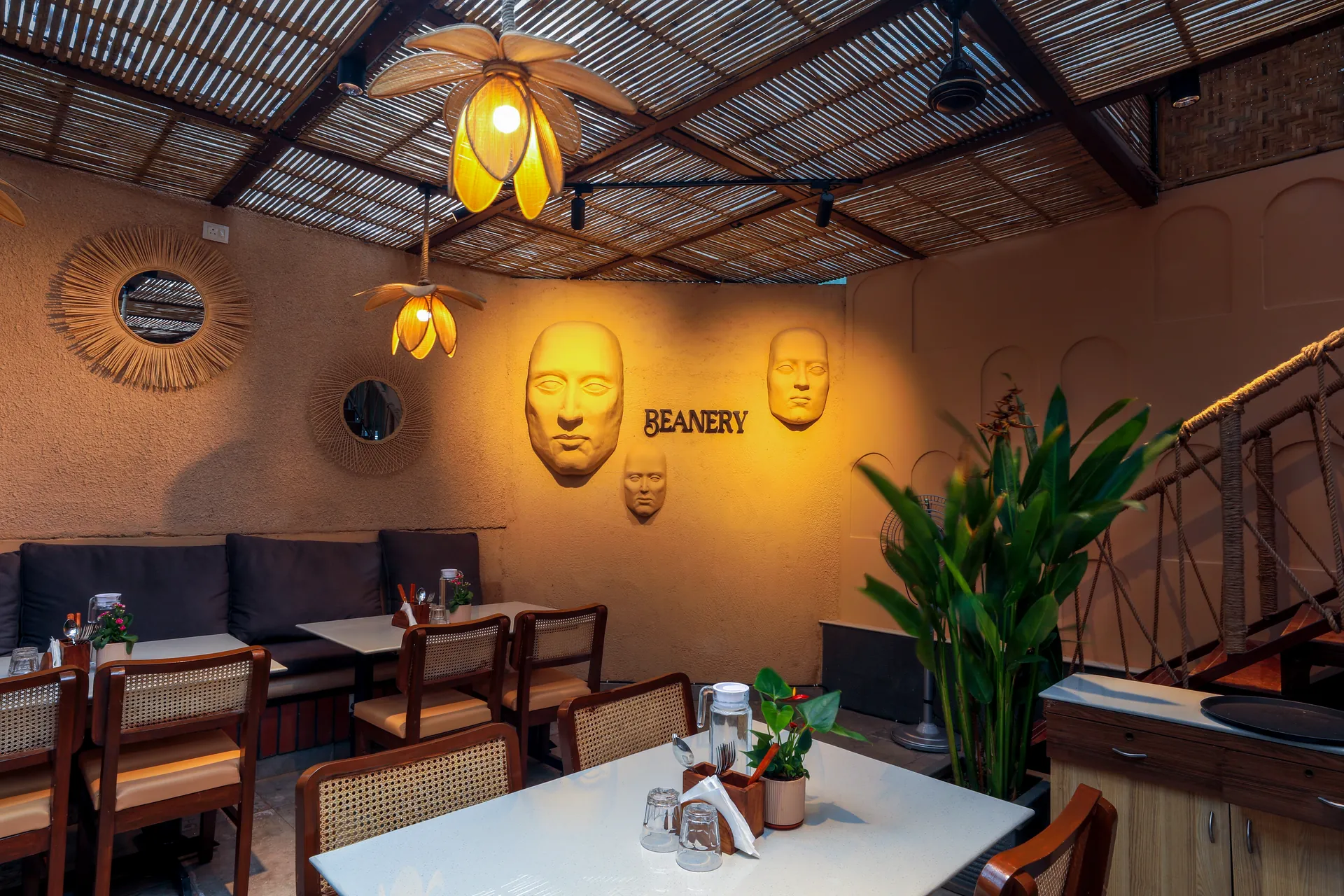 Beanery Café interior with a woven ceiling, warm petal-shaped pendant lights, cane chairs, greenery, sculpted wall faces and BEANERY signage.