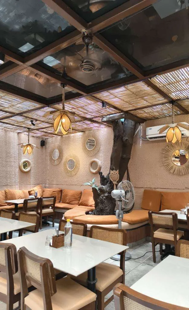 Beanery Café dining tables, cane-back chairs and orange banquette beneath the canopy beside a tree trunk.