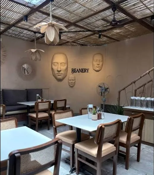 Beanery Café dining area beneath woven pendants, with sculpted-face wall artwork and the café logo.
