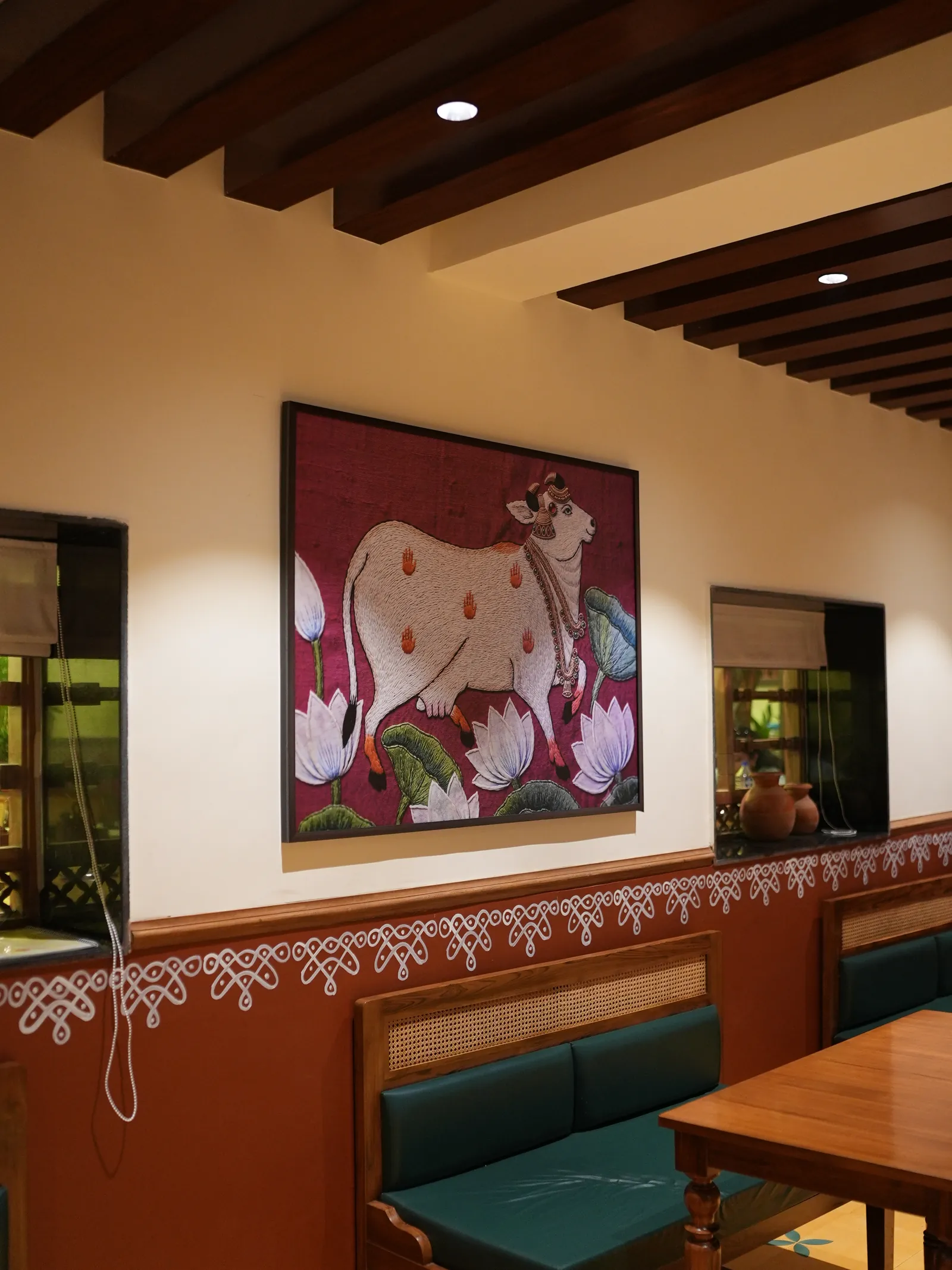 Green dining banquette below cow-and-lotus artwork, a patterned wall border and ceiling beams at Secret Sambar.